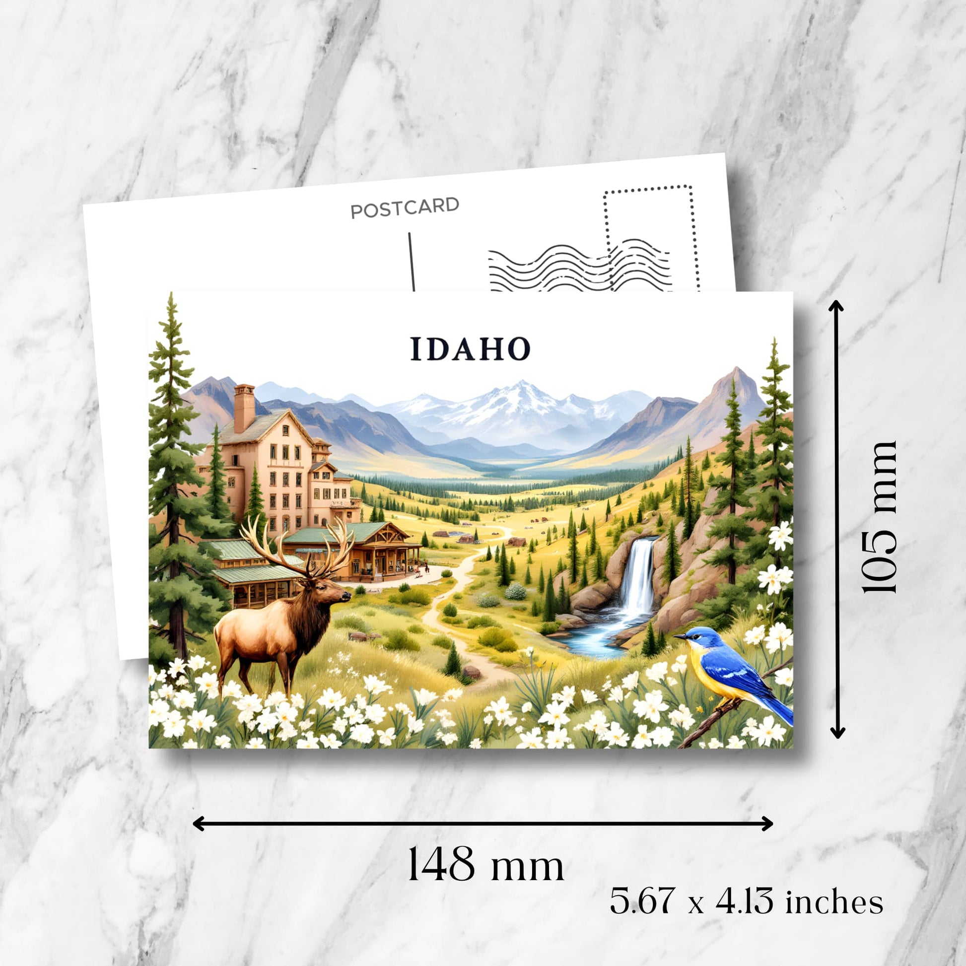 Product dimensions 148x105mm shown on Idaho souvenir postcard featuring Victorian lodge, mountain valley, elk, waterfall, and spring wildflowers framing scenic wilderness