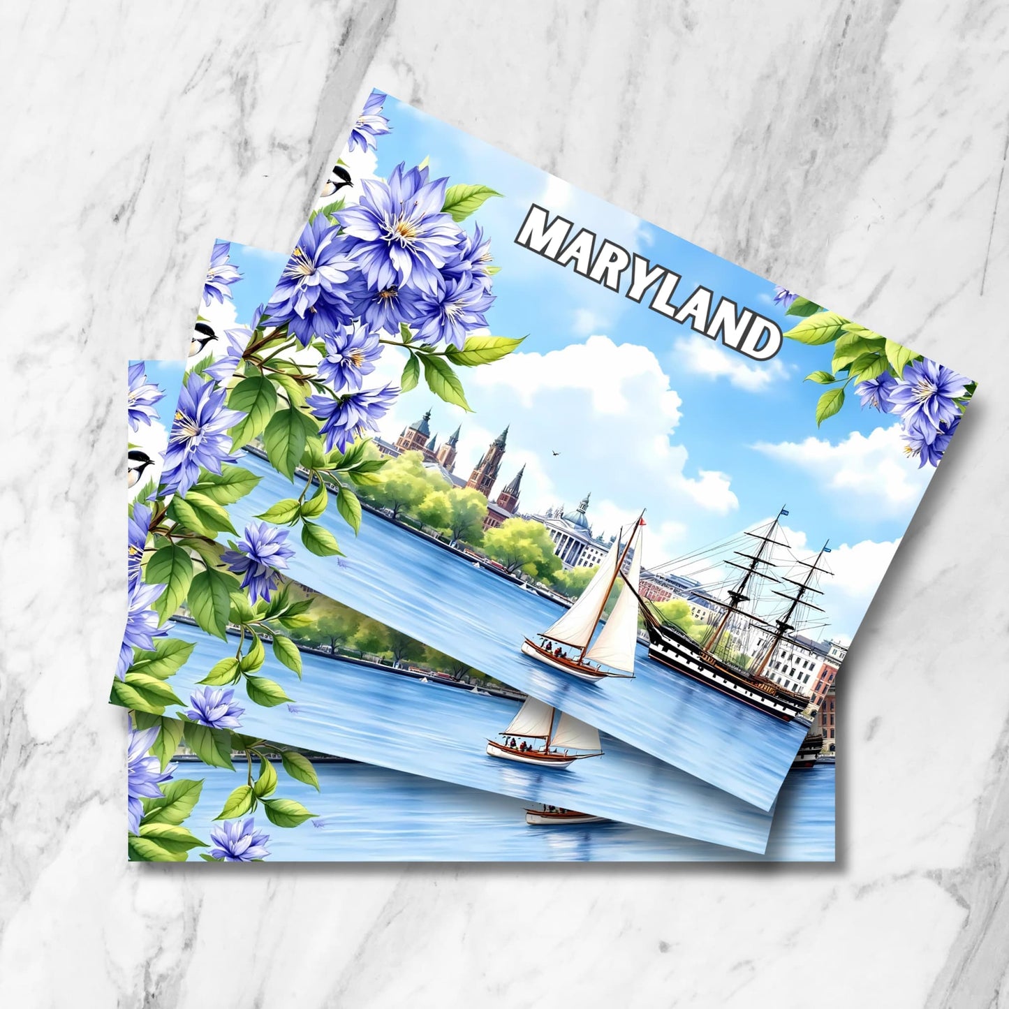 Stack of Maryland harbor prints with blue clematis flowers, sailboat, tall ship and Annapolis skyline on marble background.