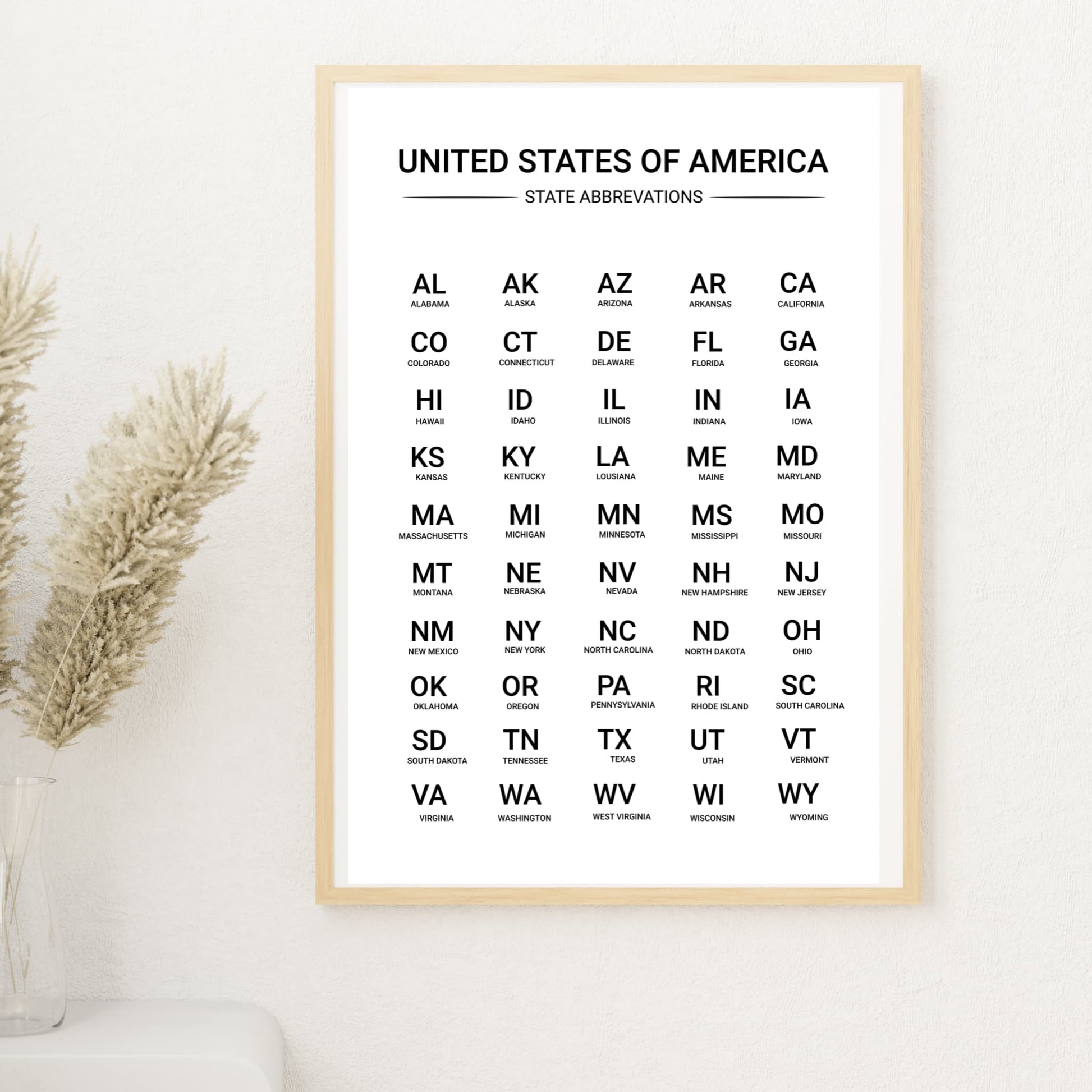 Minimalist US state abbreviations wall art framed in light wood, styled in a soft neutral interior with pampas grass and white wall background
