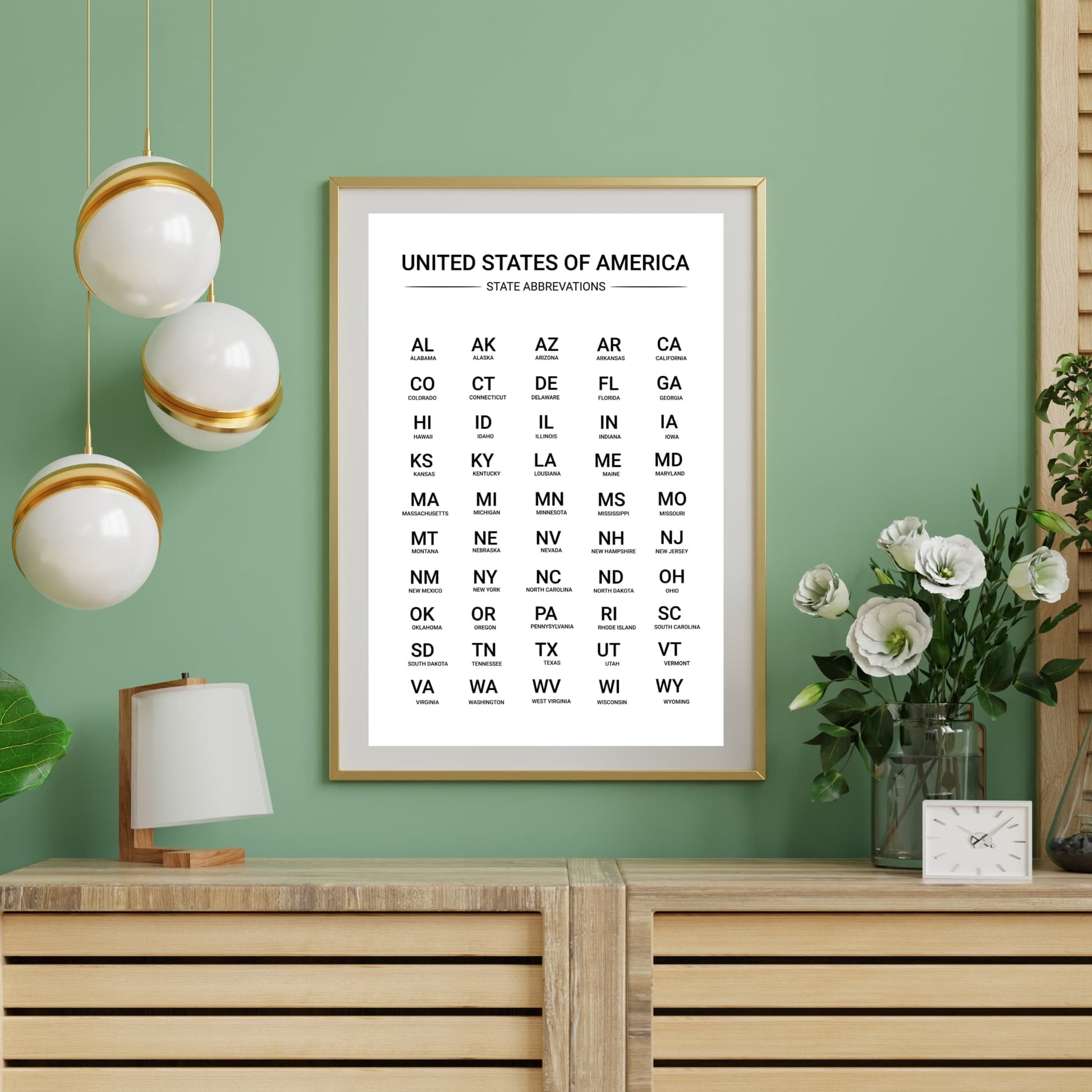 Printable US state abbreviation chart in a gold frame against a mint green wall with hanging lights and white flowers, modern home office decor