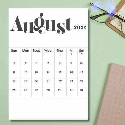 2024 August printed calendar sheet lying on top of green table with notebook and glasses besides it.