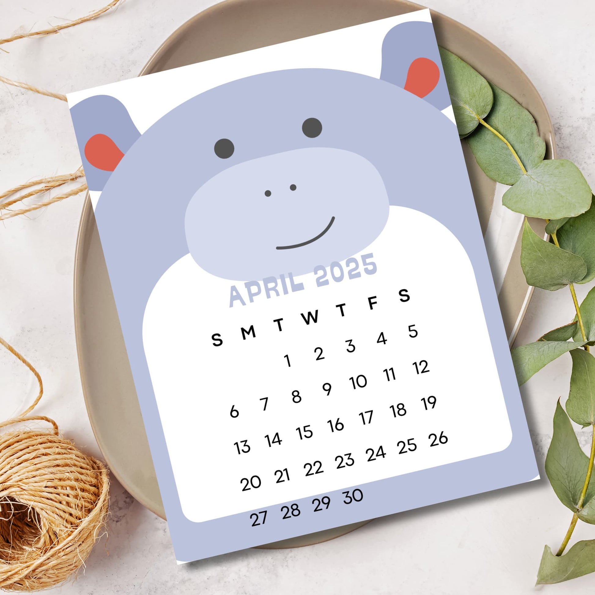 April 2025 calendar with blue hippo character. Sunday-start grid layout displayed on ceramic plate with eucalyptus leaves and natural rope elements.