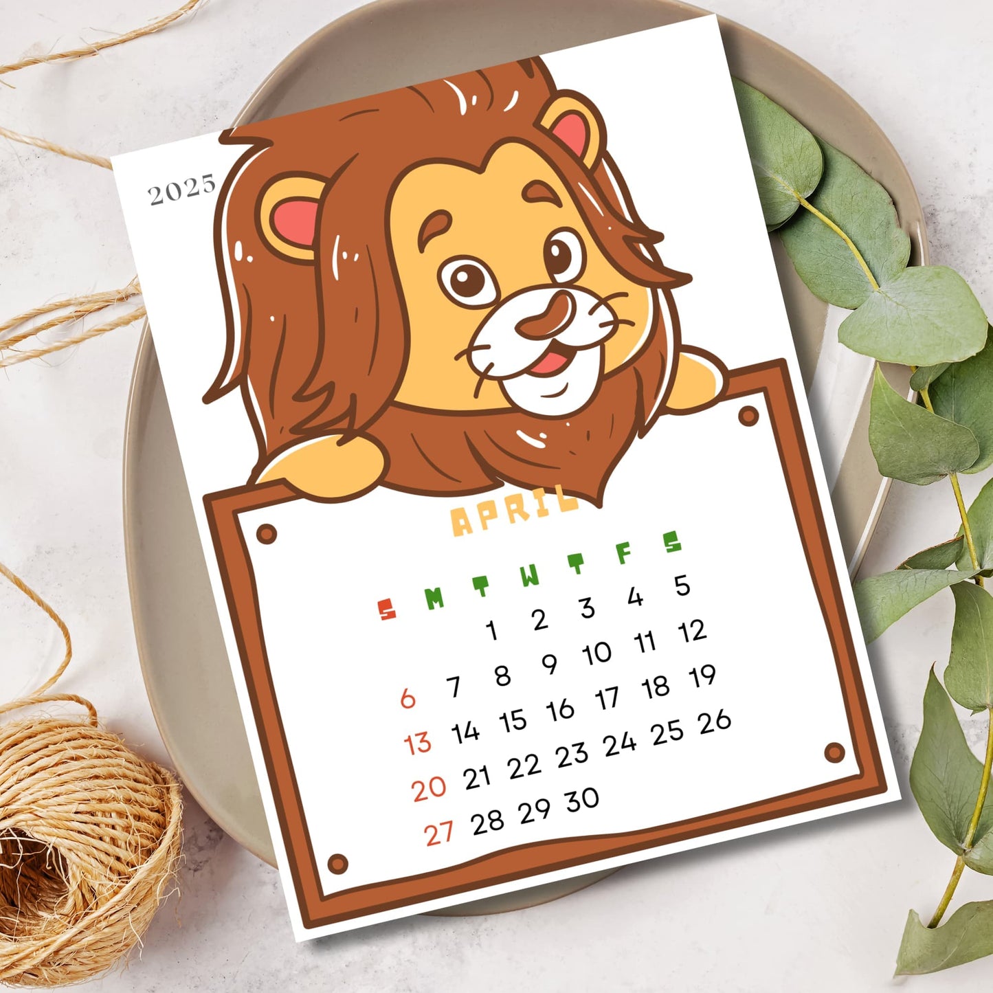 April 2025 animal calendar featuring smiling lion character. Sunday start grid with weekdays in green and weekends in red, displayed on ceramic plate with eucalyptus leaves.