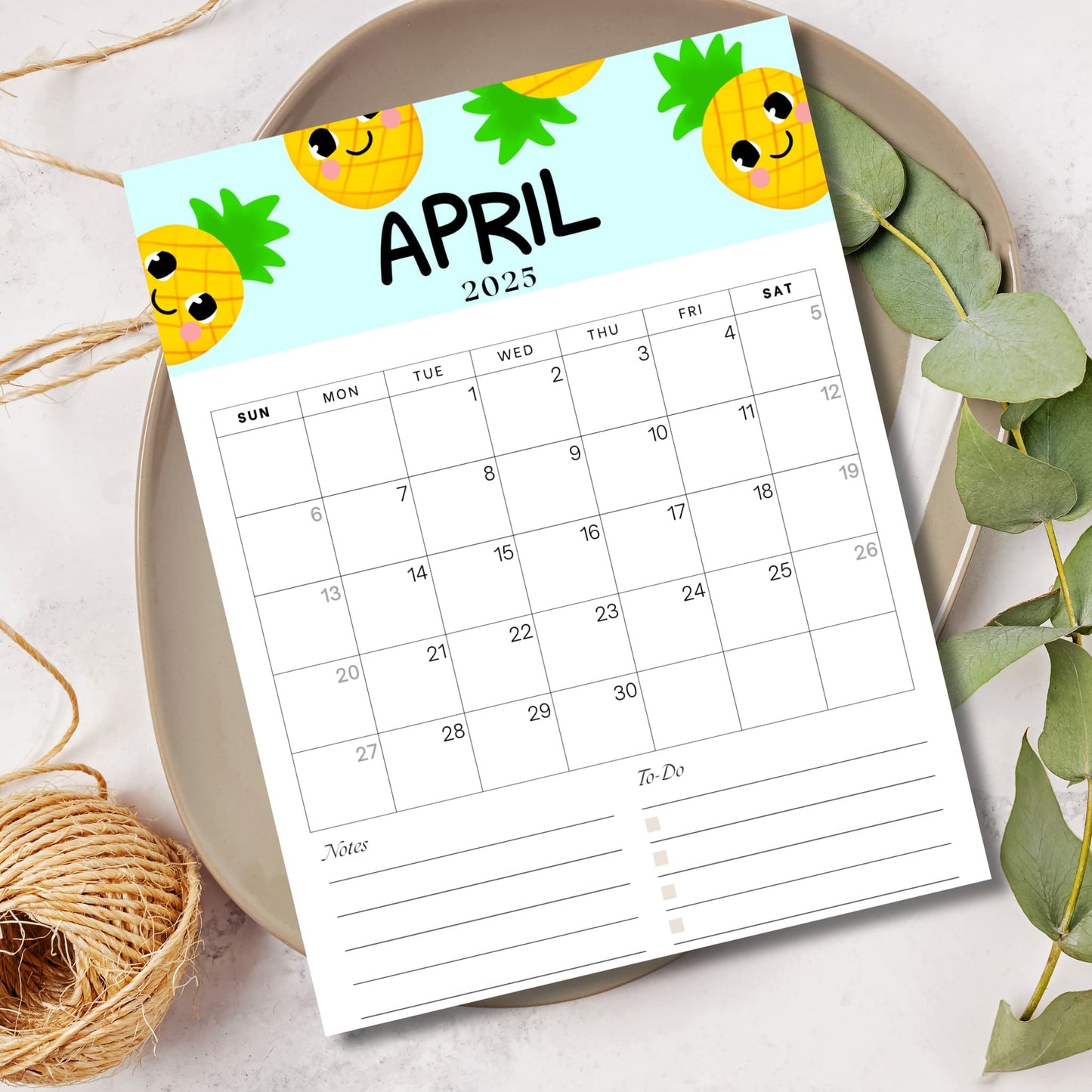 April 2025 kids calendar with smiling pineapple characters on light blue background. Sunday-start monthly planner displayed on ceramic plate with eucalyptus leaves and natural elements, ideal for homeschool planning.