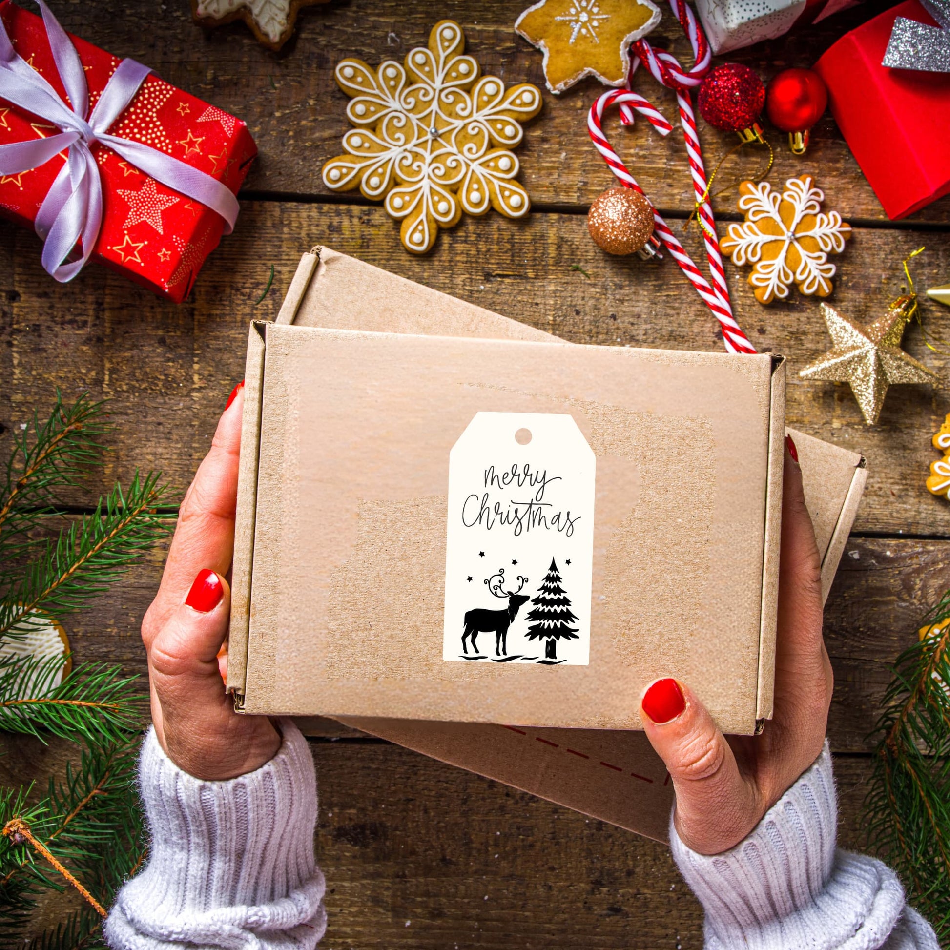 Hands holding kraft box decorated with white Christmas gift tag featuring reindeer and pine tree design, surrounded by gingerbread cookies, candy canes, and holiday decorations on rustic wooden surface