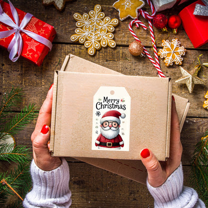 Hands holding kraft gift box decorated with cute Santa gift tag wearing glasses, styled with gingerbread cookies, candy canes, and pine branches on rustic wooden surface