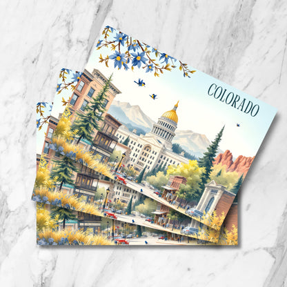 Vintage-style Denver cityscape prints featuring State Capitol with golden dome, historic buildings, Rocky Mountains backdrop, and state flower blue columbines, shown as stacked postcards on marble background