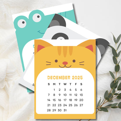 December 2025 calendar with yellow cat design, featuring frog and panda pages behind. Sunday-start grid layout with eucalyptus sprig decoration.