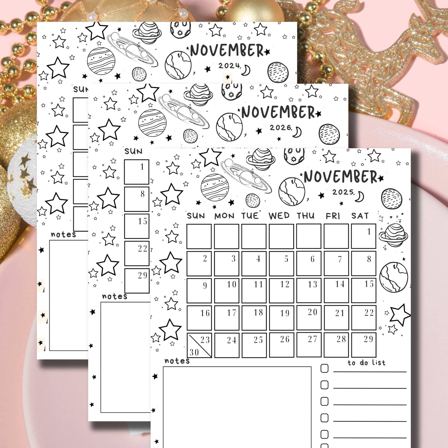 Layered November calendars with celestial doodles and planets, black and white design, displayed with golden holiday decorations on pink