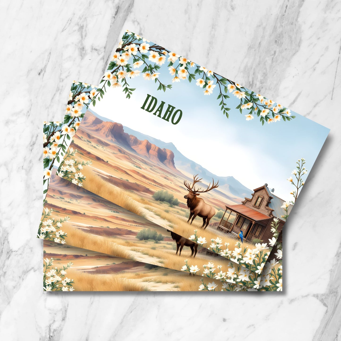 Stacked vintage-style Idaho postcards featuring desert mountain landscape, majestic wild elk, rustic wooden cabin, and decorative spring blossoms framing scenic western vista