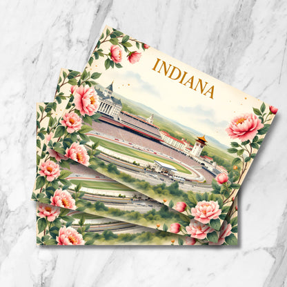 Artistic Indiana travel postcard featuring Indianapolis Motor Speedway surrounded by pink peonies, with skyline backdrop, red cardinals, and golden dome statehouse. Multiple cards shown stacked on marble surface.