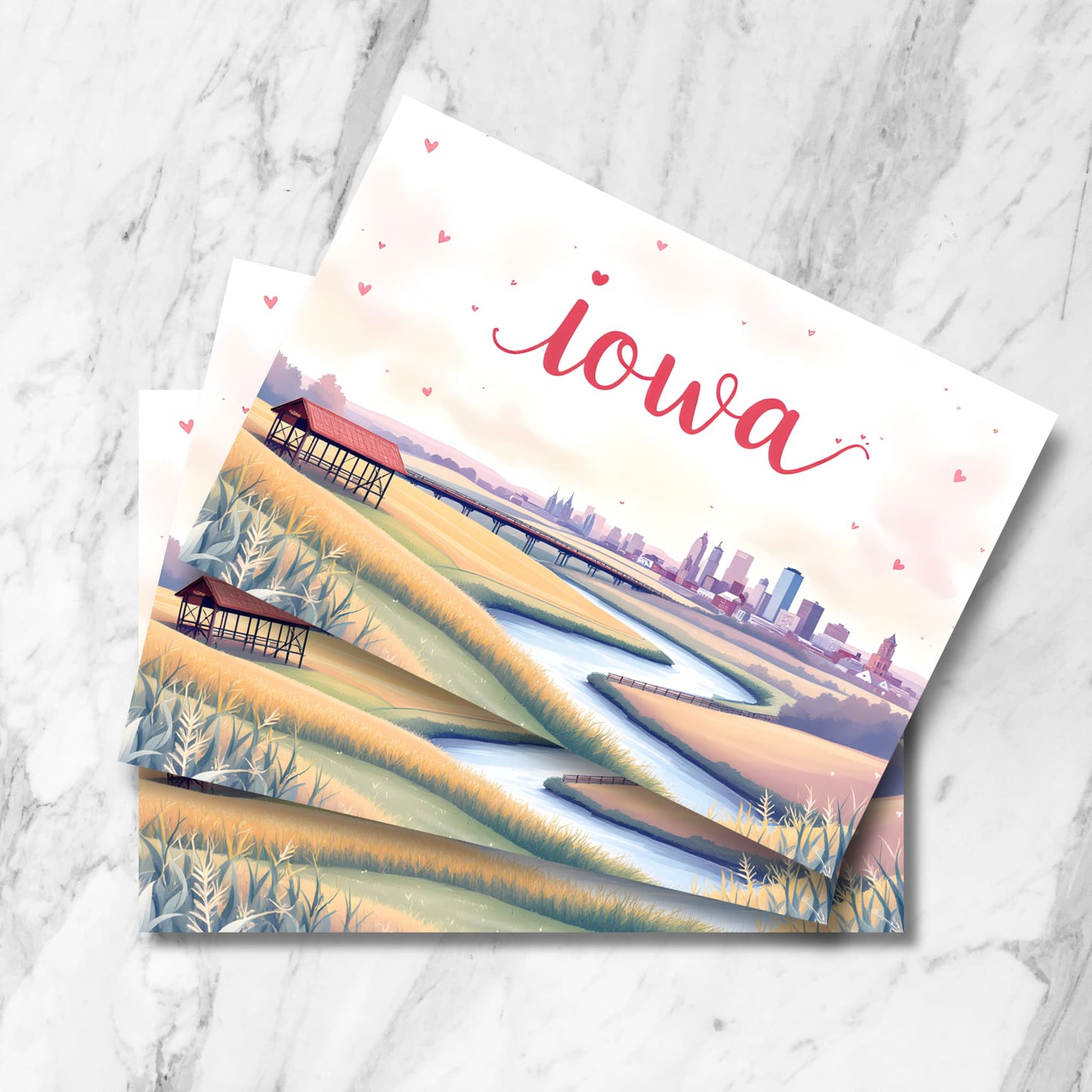 Romantic Iowa postcard featuring Des Moines skyline at sunset with meandering river, wheat fields, and rustic covered shelter. Illustrated in soft pastel colors with floating heart details and handlettered state name.