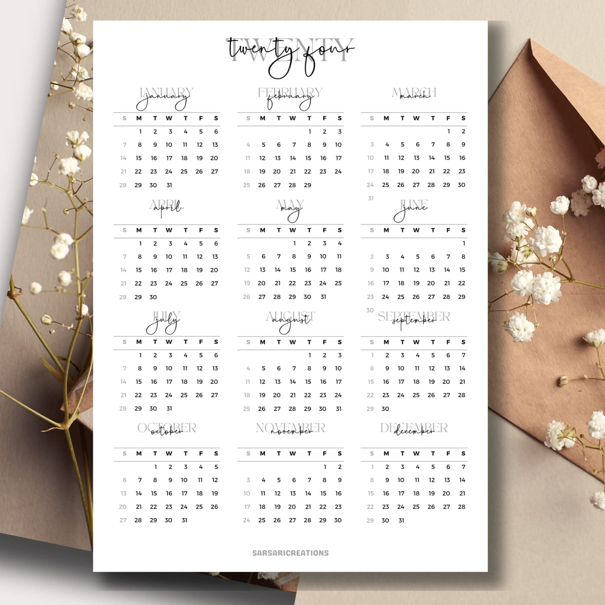 Script font style 2024 full year calendar page on a table with envelope and small white flowers in the background.