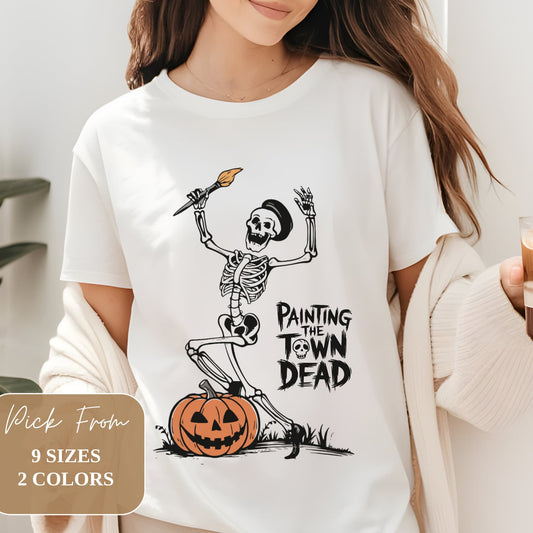 White t-shirt featuring skeleton painter with paintbrush standing on pumpkin. Text reads 'Painting the Town Dead'. Available in 9 sizes, 2 colors.