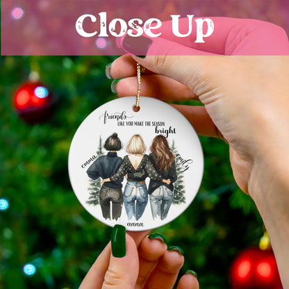 Close-up of personalized best friends ornament held against lit Christmas tree, showing detailed illustration of three friends in winter outfits with customizable names Emma, Anna, and Emily.